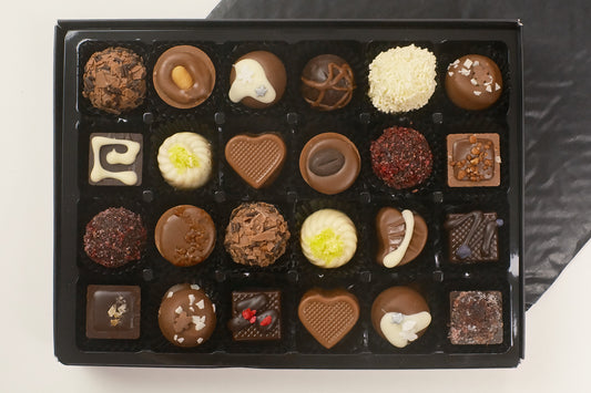 A black gift box filled with 24 assorted chocolates, including heart-shaped, round, and square pieces in various chocolate tones and toppings, photographed from above on a light surface with a dark cloth in the background.