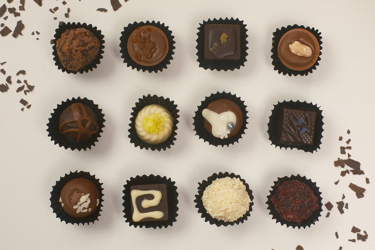 A top-down arrangement of twelve assorted chocolate truffles in black paper cups on a light beige background with scattered chocolate shavings.