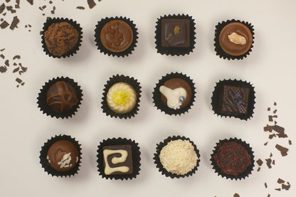 A top-down arrangement of twelve assorted chocolate truffles in black paper cups on a light beige background with scattered chocolate shavings.