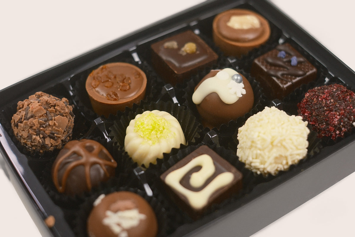 A black gift box filled with assorted gourmet chocolates, including milk and dark chocolate pieces, white chocolate truffles, and decorated squares, photographed on a light neutral background.