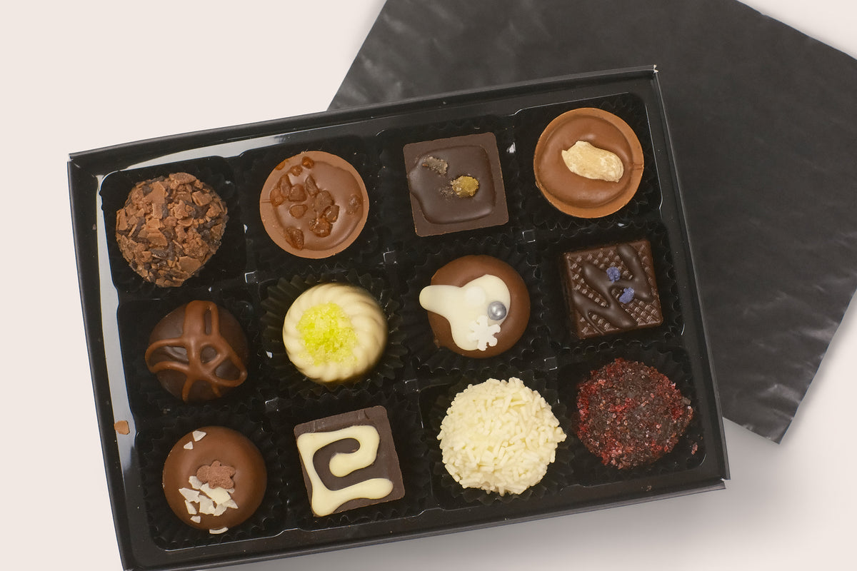 An assortment of gourmet chocolates in a black gift box with the lid placed behind it on a light background.