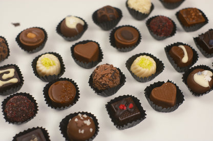 An assortment of 24 gourmet chocolates in black paper cups, including milk and dark chocolate pieces with white chocolate drizzle, sprinkles, and heart-shaped molds, arranged on a white background.