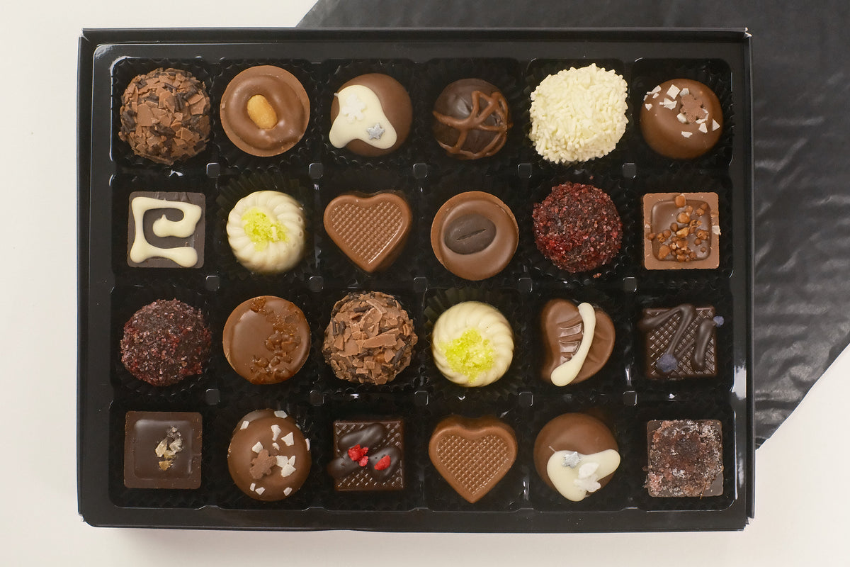 A black gift box filled with 24 assorted chocolates, including heart-shaped, round, and square pieces in various chocolate tones and toppings, photographed from above on a light surface with a dark cloth in the background.