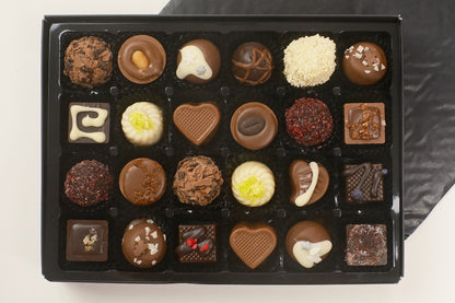 A black gift box filled with 24 assorted chocolates, including heart-shaped, round, and square pieces in various chocolate tones and toppings, photographed from above on a light surface with a dark cloth in the background.