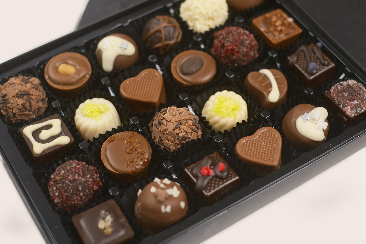 A black gift box filled with assorted chocolates, including heart-shaped, round, and square pieces in dark, milk, and white chocolate with various toppings, photographed on a light background.