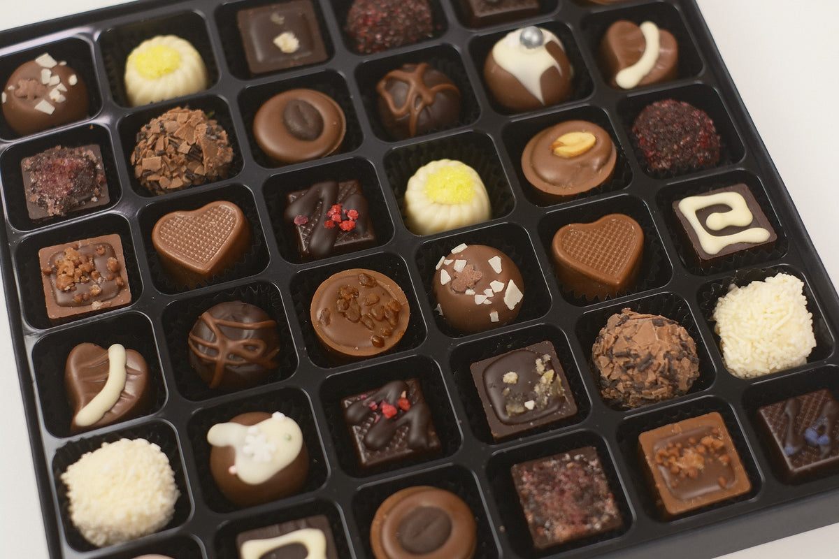 A black tray filled with 36 assorted chocolates in various shapes and decorations, photographed from above on a light background.