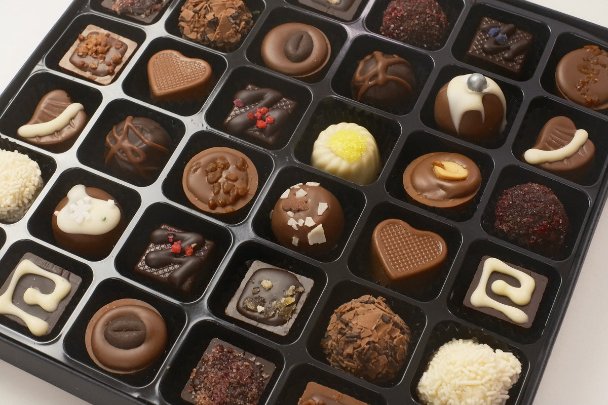 A black chocolate box filled with 36 assorted gourmet chocolates, truffles, and pralines in various shapes and flavors, photographed from a high angle on a light background.