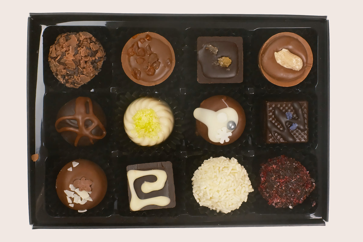 A 12-piece assortment of gourmet chocolates in a black gift box, featuring various shapes, textures, and toppings like white chocolate, caramel, and nuts, photographed from directly above on a light beige background.