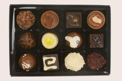 A 12-piece assortment of gourmet chocolates in a black gift box, featuring various shapes, textures, and toppings like white chocolate, caramel, and nuts, photographed from directly above on a light beige background.