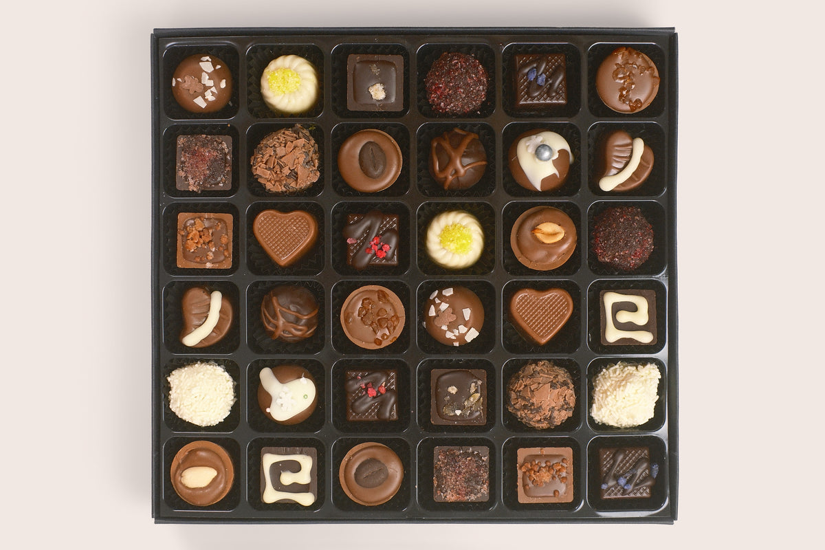 A black chocolate box filled with assorted gourmet chocolates, including various shapes, sizes, and decorations like nuts, sprinkles, and white chocolate drizzle, arranged in a neat grid on a light beige background.