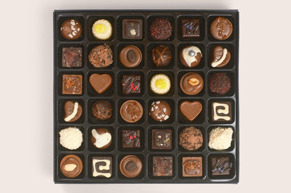 A black chocolate box filled with assorted gourmet chocolates, including various shapes, sizes, and decorations like nuts, sprinkles, and white chocolate drizzle, arranged in a neat grid on a light beige background.