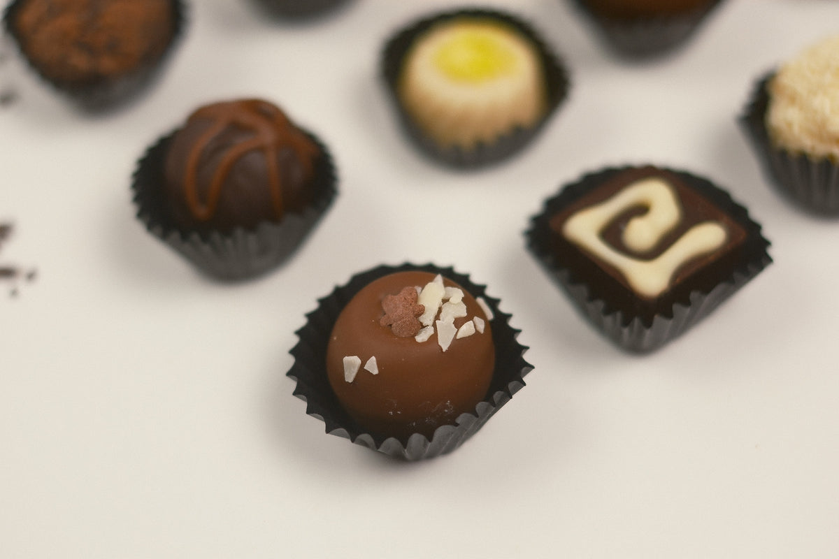 A close-up, shallow depth-of-field shot of assorted gourmet chocolate truffles in black paper cups on a clean white surface. The central, sharpest truffle is coated in milk chocolate and topped with white chocolate shavings, surrounded by other chocolate truffles in soft focus.