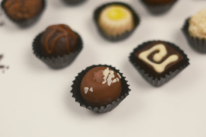 A close-up, shallow depth-of-field shot of assorted gourmet chocolate truffles in black paper cups on a clean white surface. The central, sharpest truffle is coated in milk chocolate and topped with white chocolate shavings, surrounded by other chocolate truffles in soft focus.