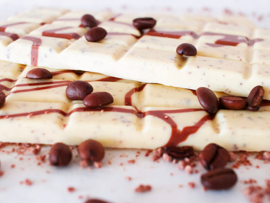Image shows a close up of 2, 100g white chocolate, coffee flavoured hand made chocolate bars, scattered with coffee beans.